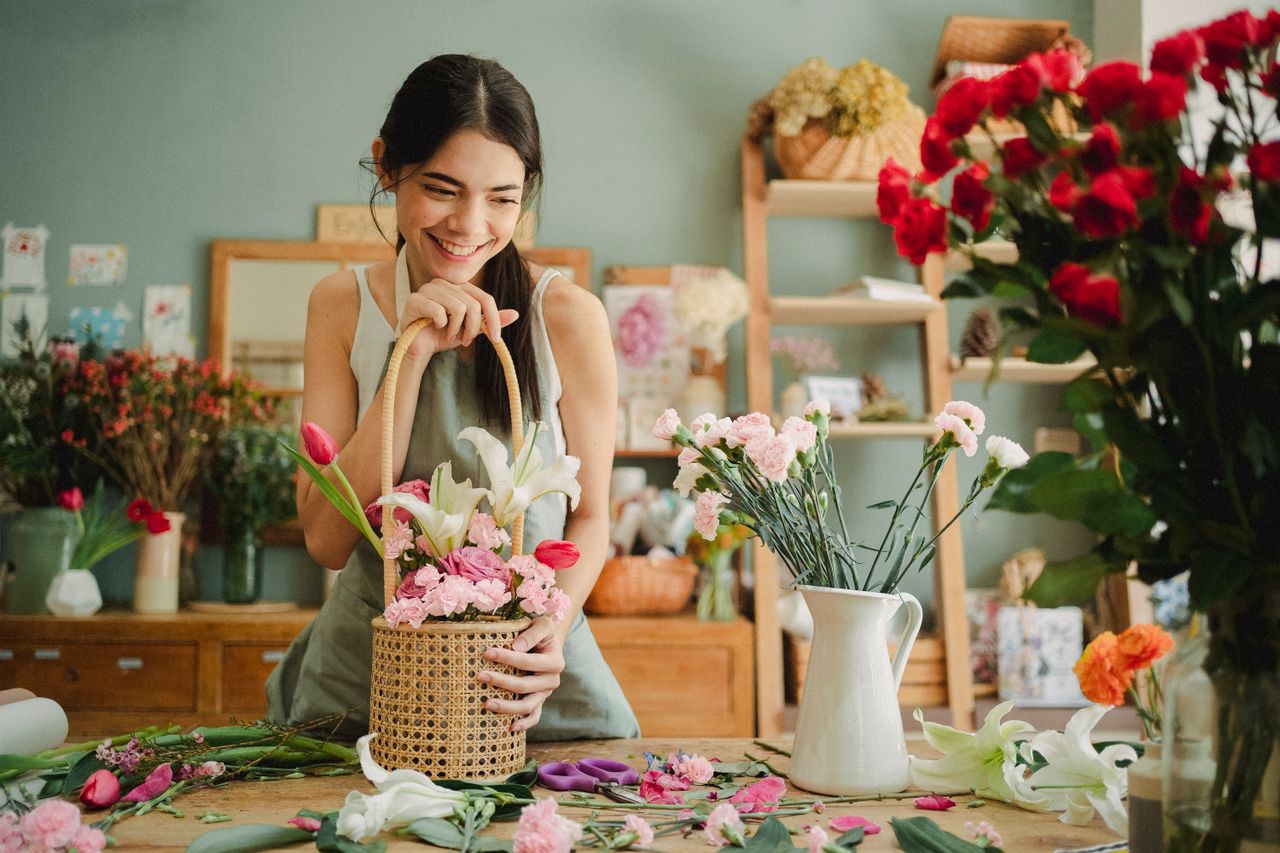 Positive ethnic florist with long dark hair in apron leaning on wicker basket while choosing flowers for composition in floristry store with cozy interior