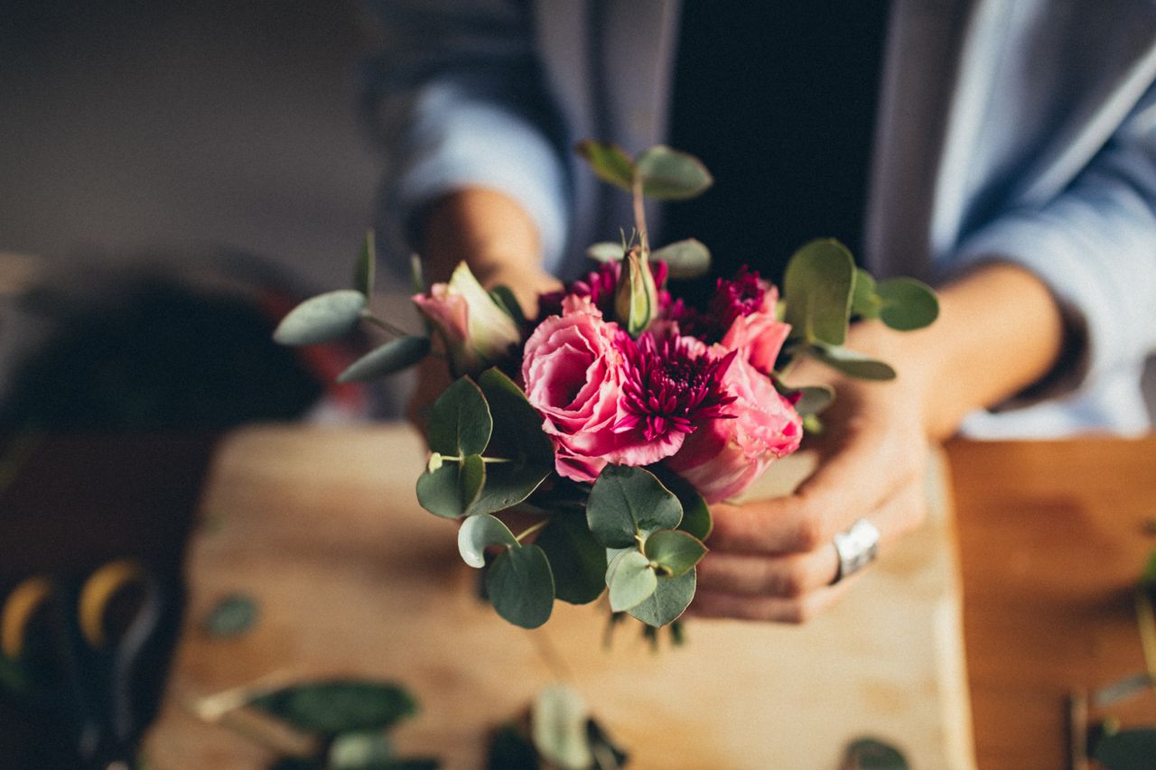 A florist arranges a vibrant bouquet of pink flowers and green leaves on a wooden table.
